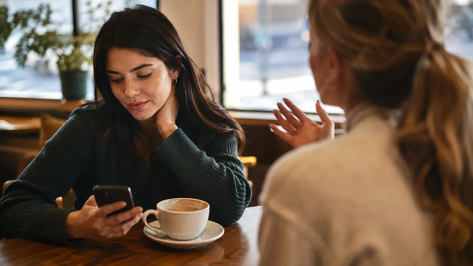 A person at a café table glancing at their phone while a friend across the table talks, coffee half finished